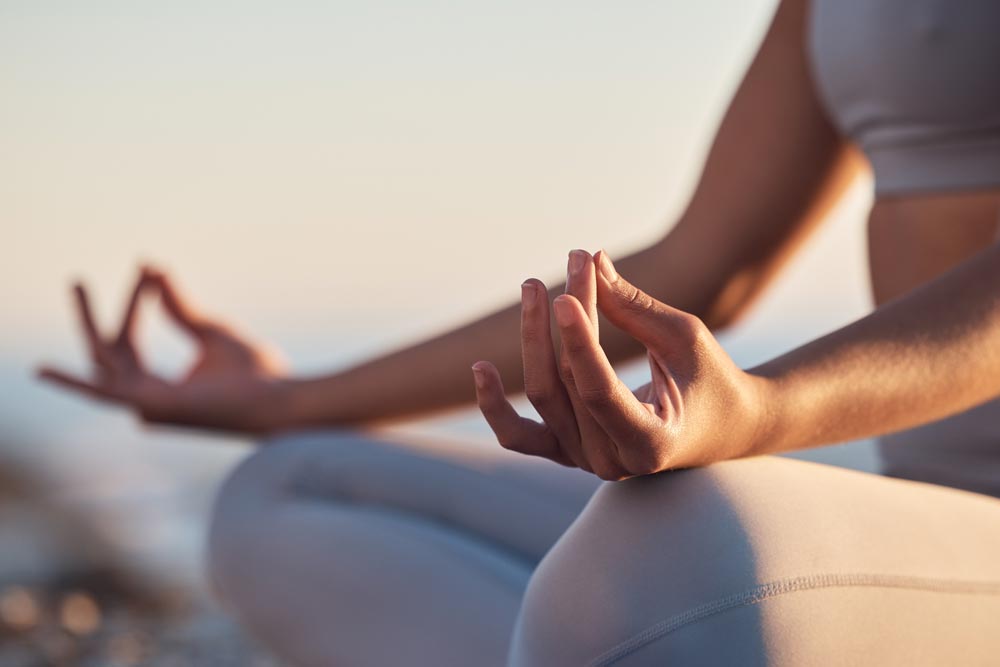 En ung kvinna sitter i lotusställning och mediterar vid strandkanten under gyllene solnedgång